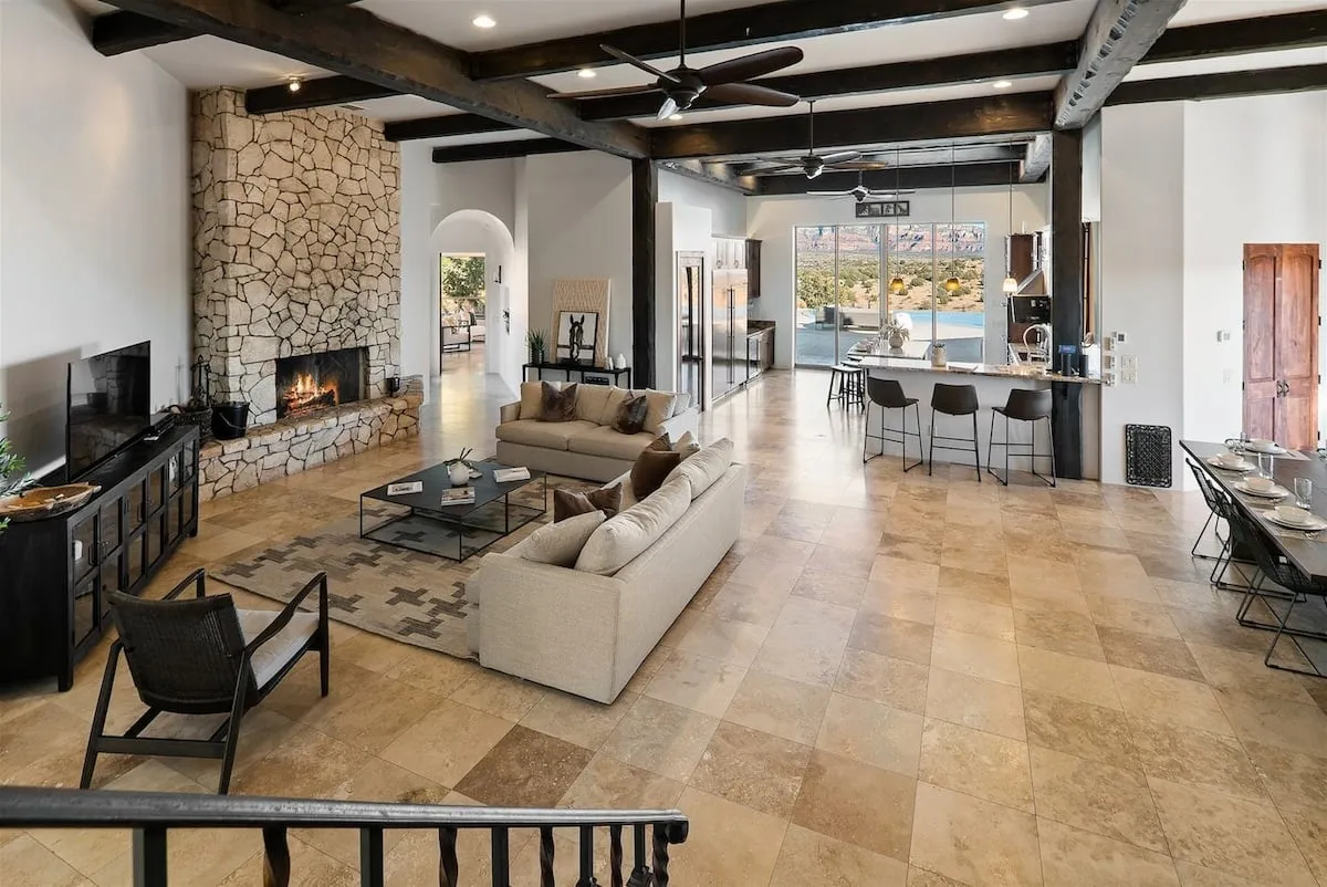 A spacious open-concept interior featuring a striking stone fireplace, comfortable seating arranged for conversation, large windows flooding the space with natural light, and a seamless transition into the gourmet kitchen area. The overall mood is warm and welcoming, ideal for group gatherings.