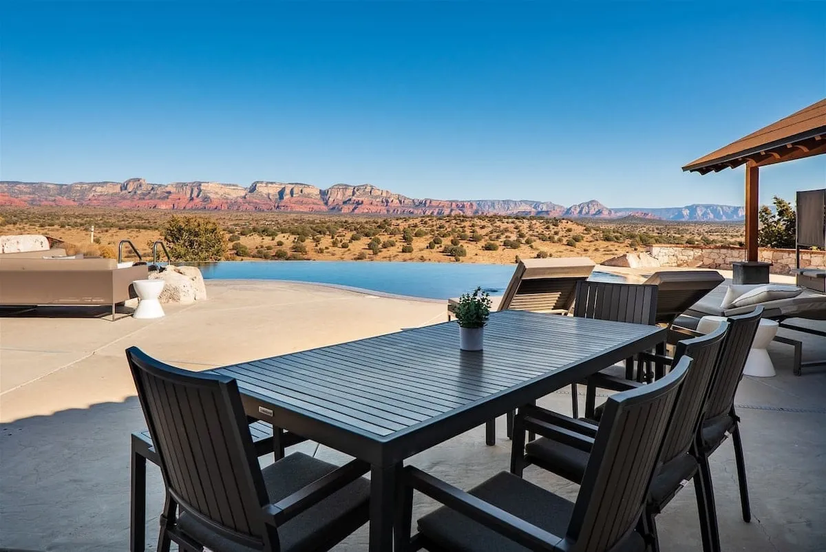 A stylish outdoor dining area overlooks an infinity pool and distant mountains, with lounge chairs nearby. The setting suggests the perfect location for alfresco meals, sunset swims, and relaxed social gatherings.