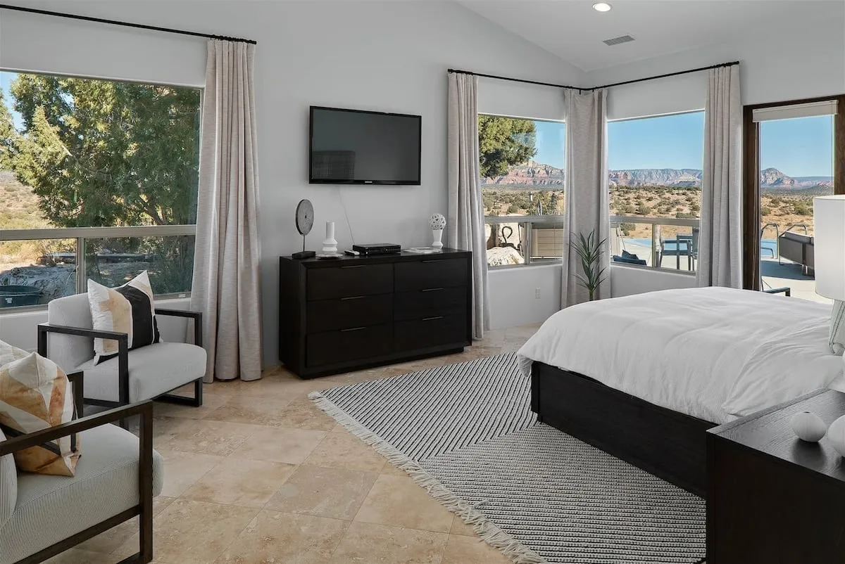 A spacious, light-filled bedroom with large windows that offer panoramic views, a comfortable bed with white linens, a modern dresser, and a cozy seating area positioned to take in the scenery. The space is styled for both relaxation and enjoying quiet moments.
