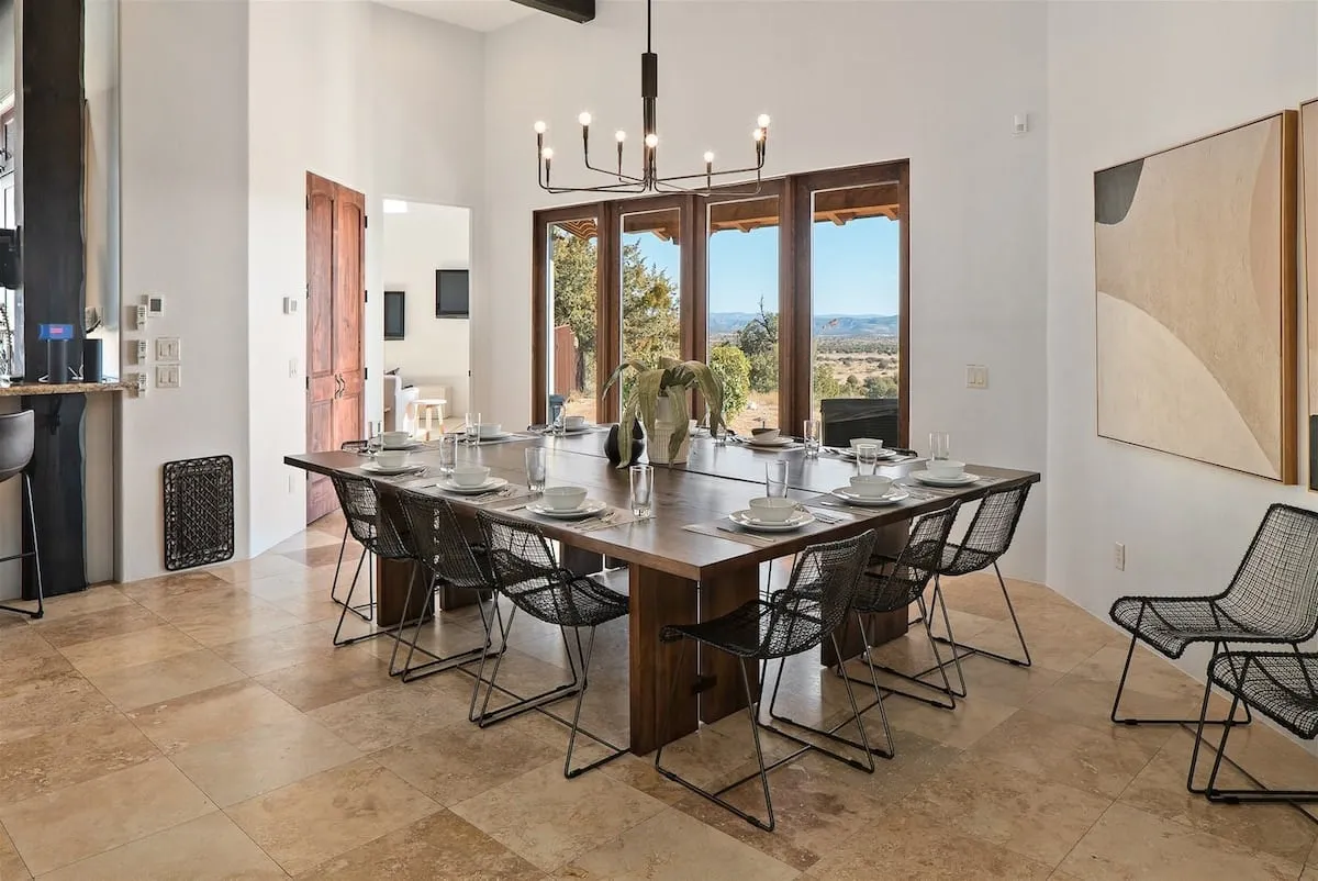 An expansive wooden dining table sits beneath a striking chandelier, surrounded by floor-to-ceiling windows that offer panoramic views. The space feels open and airy, making it perfect for large group dinners or intimate morning coffees while taking in the scenery.