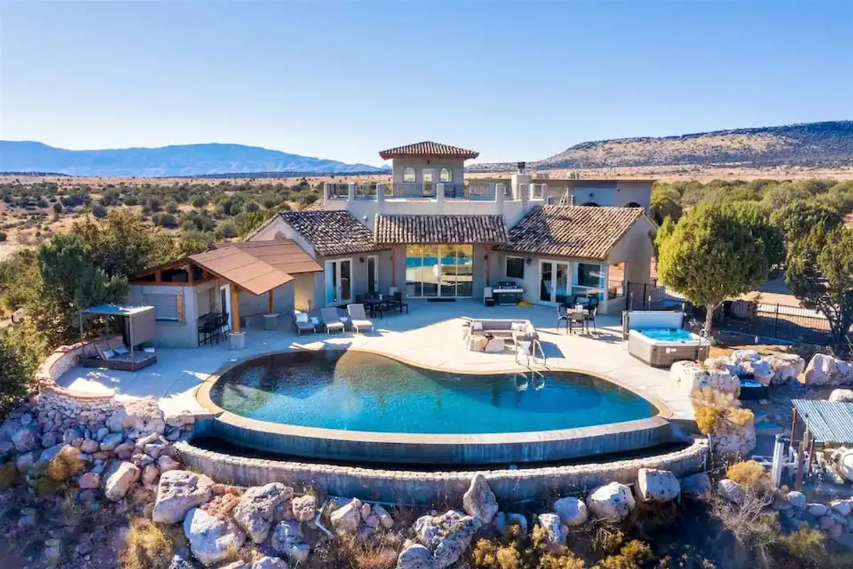 The image showcases a luxurious infinity pool overlooking Sedona’s iconic Red Rocks, with a spacious patio perfect for outdoor dining and elegant Southwestern architecture. The golden sunset casts a warm glow, inviting guests to relax by the pool or gather around the fire pit.