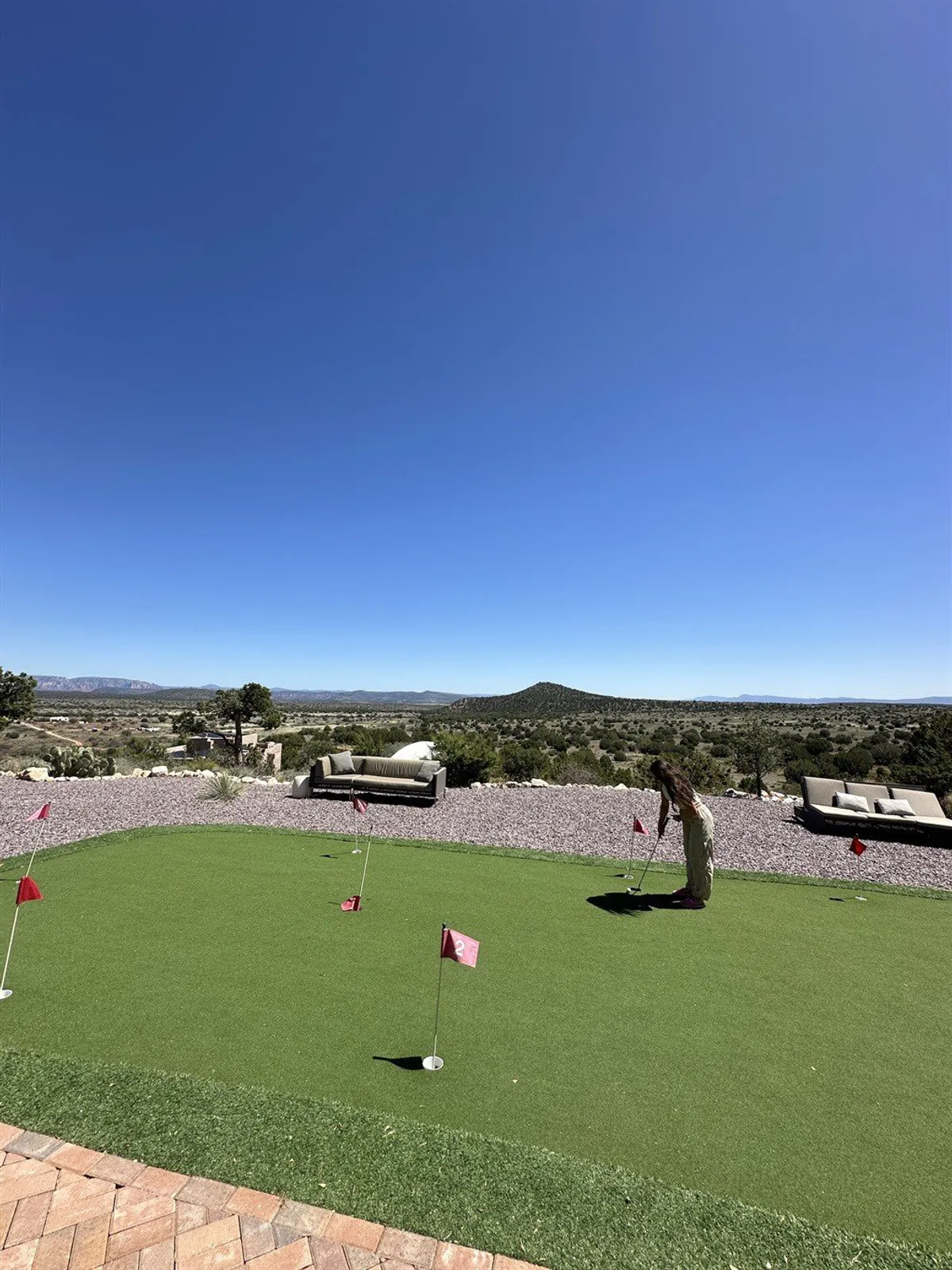 The image features a lush putting green with red flags, comfortable lounge seating, and a dramatic mountain view. The setting is playful yet serene, ideal for friendly golf competitions or casual relaxation.