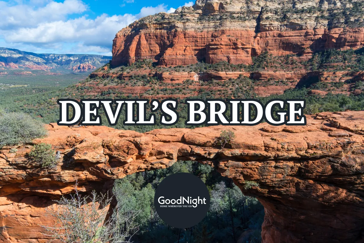 This image showcases Devil's Bridge, a dramatic red rock formation set among rugged terrain and lush green vegetation, with distant mountains visible under an expansive sky. The scene is vibrant and adventurous, capturing the iconic landscape that makes Sedona famous for hiking and exploration.