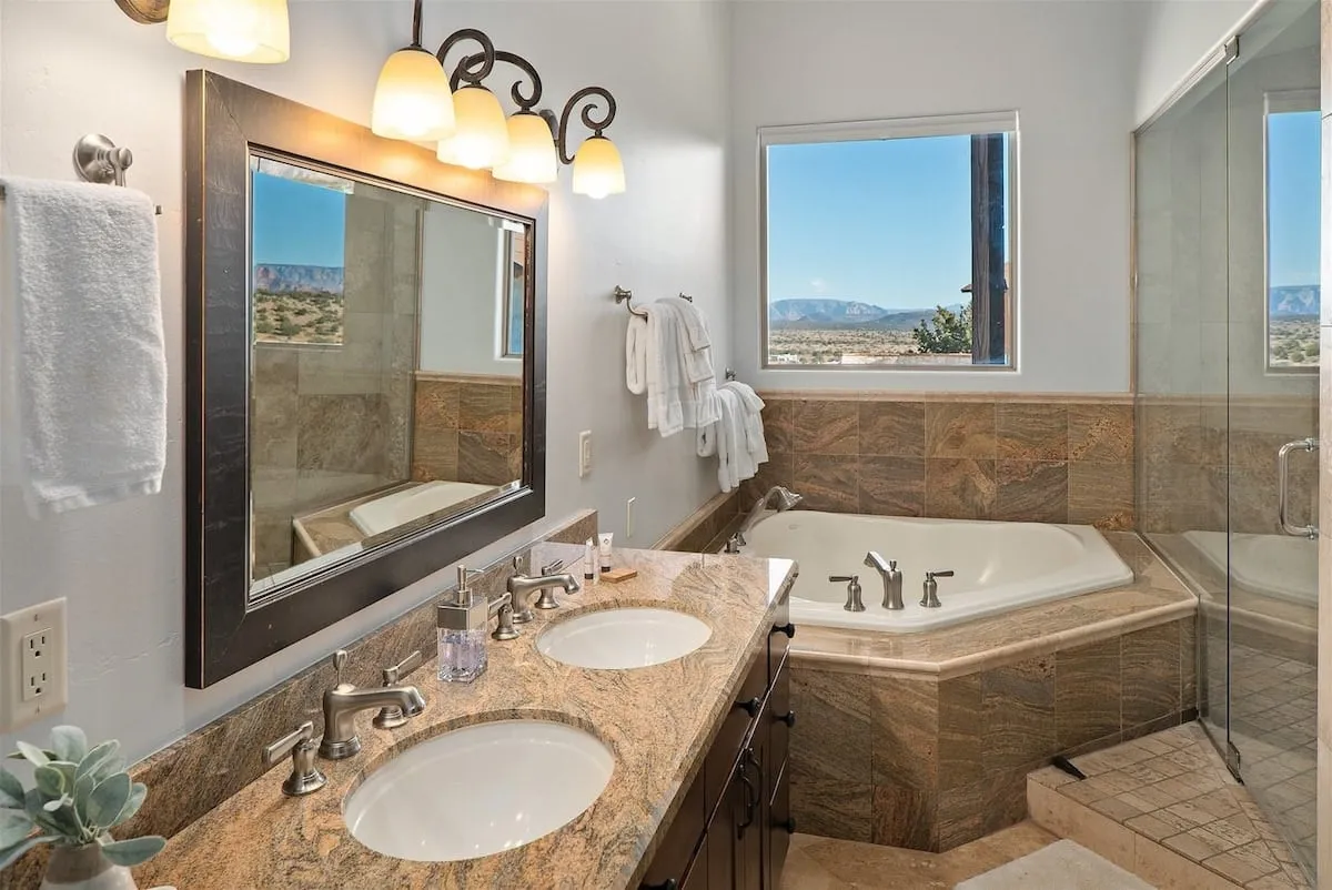 A luxurious bathroom featuring a double vanity sink, a spacious soaking tub set beside a window with sweeping mountain views, and a mirror illuminated by warm, ambient lighting. The materials and design evoke a spa-like atmosphere, perfect for relaxation and self-care.