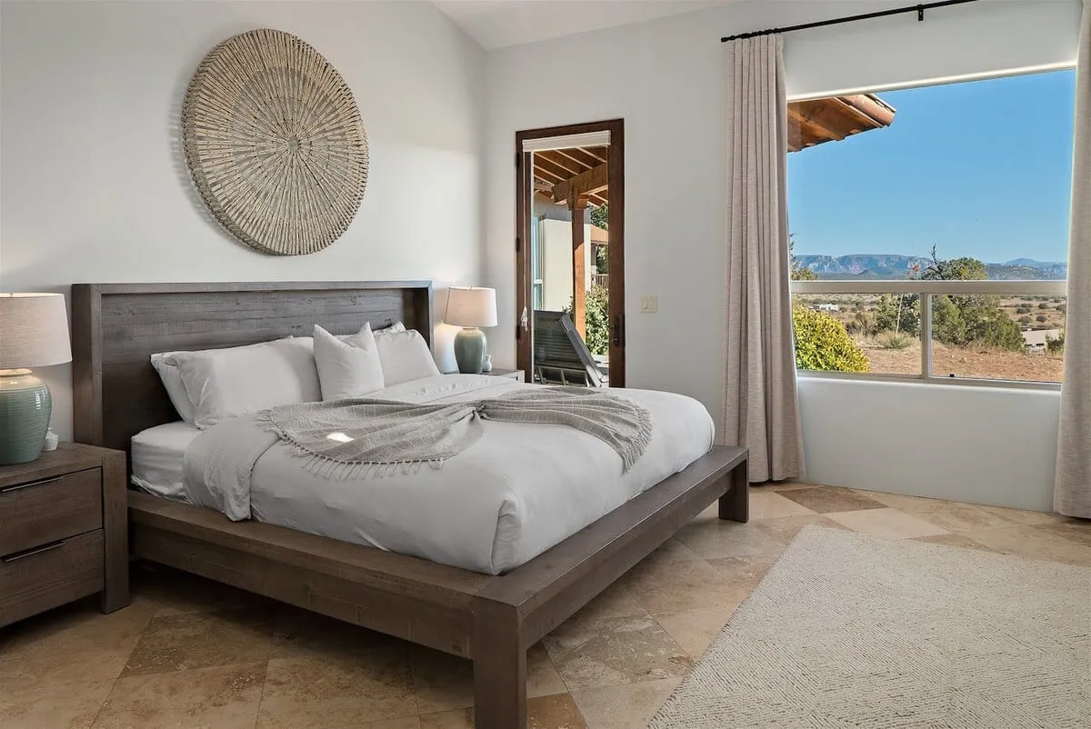 A tranquil bedroom scene featuring a bed with crisp white linens, a decorative wall piece above the headboard, and a large window that frames a beautiful mountain view. The room exudes calm with soft lighting and minimalistic décor, emphasizing comfort and relaxation.