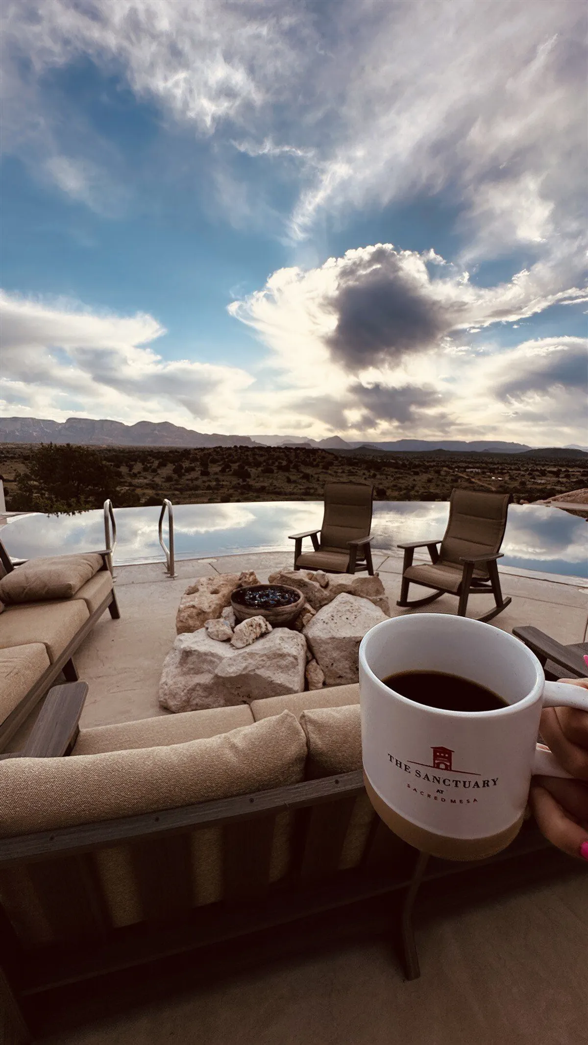 This image captures an expansive infinity pool set against a breathtaking mountain backdrop, with comfortable lounge chairs, a cozy fire pit, and a coffee cup on the deck. The scene is tranquil and inviting, showcasing both water and fire features, with the pool water reflecting the sunset.