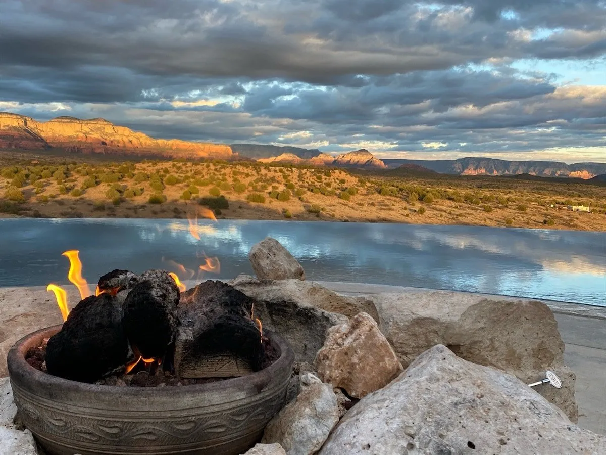A cozy fire pit sits overlooking distant mountains and shimmering water, inviting groups to gather around as the sun sets. The serene environment is perfect for intimate conversations and stargazing.