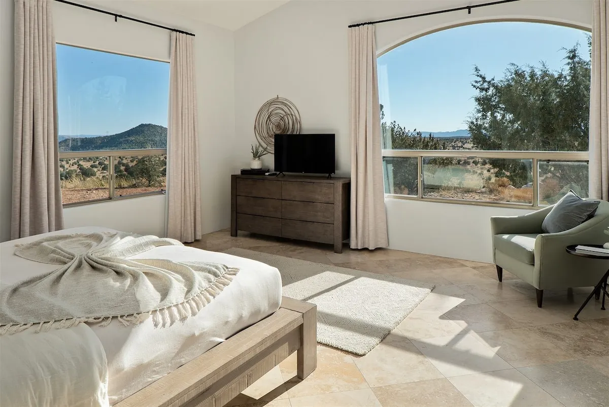 A cozy bedroom scene with a decorative throw blanket on the bed, a TV atop a modern dresser, a green accent chair, and large windows that open to a scenic mountain landscape. The room offers both comfort and entertainment, making it ideal for family or group stays.